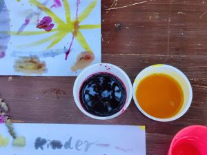 Zwei Becher mit Pflanzenfarben – kräftiges Rot und leuchtendes Gelb – neben einem mit Naturfarben gemalten Kinderbild.