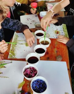 Kinder malen mit Pflanzenfarben am Tisch; verschiedene Blüten und Farbtöpfchen stehen bereit.