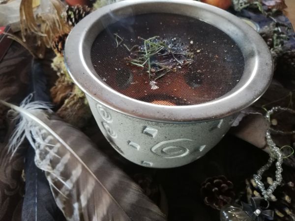 Räucherstövchen mit glimmendem Räucherwerk auf Sieb, umgeben von Naturmaterialien wie Feder, Zapfen und Moos – herbstlich-mystische Stimmung passend zum Jahreskreisfest Samhain.