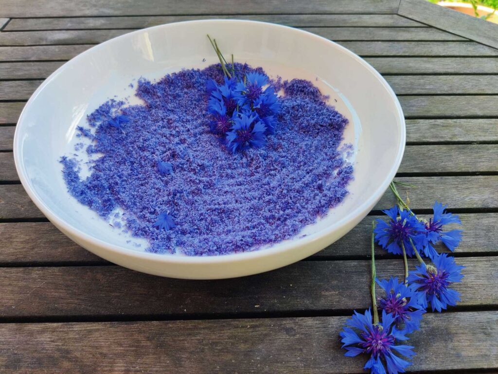 Leuchtend blaues Kornblumen-Blütensalz in einer weißen Schale; frische Kornblumen liegen daneben auf dem Holztisch.