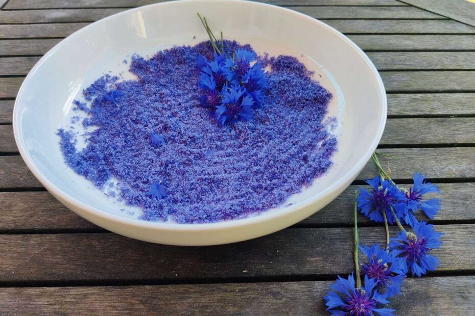 Leuchtend blaues Kornblumen-Blütensalz in einer weißen Schale; frische Kornblumen liegen daneben auf dem Holztisch.