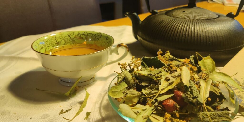 Tasse Immunfit-Tee neben einer schwarzen Teekanne und getrockneten Lindenblüten, Hagebutten und Kräutern auf dem Tisch.