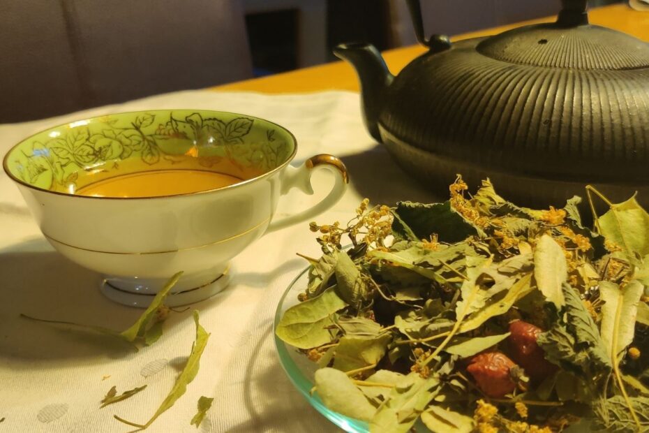 Tasse Immunfit-Tee neben einer schwarzen Teekanne und getrockneten Lindenblüten, Hagebutten und Kräutern auf dem Tisch.